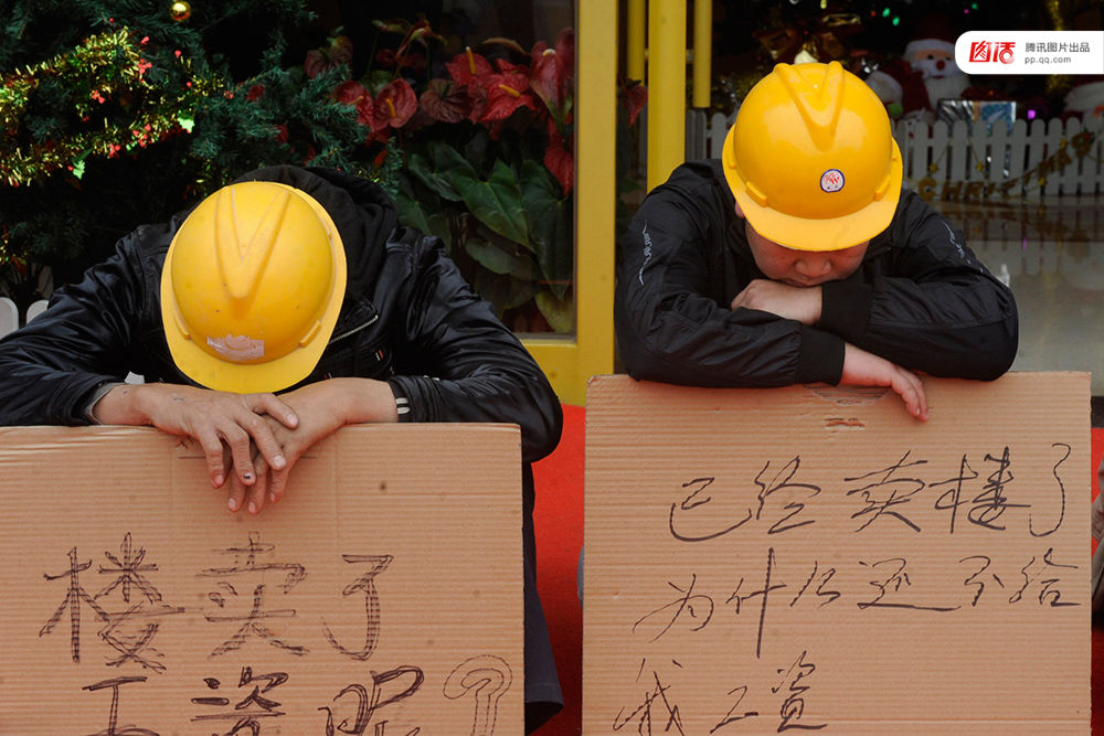 2012年12月25日，广东中山，火炬开发区的多名建筑工人讨要工作一年多的血汗钱一筹莫展，一些工人干脆将铺盖卷搬到了售楼部，并在门口搭起灶台煮饭，希望以这种方式讨回工钱。图为在销售中心门外，工人举牌讨薪。吴进/视觉中国